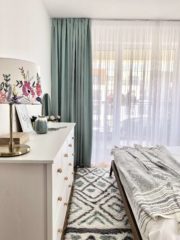В спалнята се появява цветен акцент в завесите и килима. Тук се повтаря концепцията за комбиниране на бяло с дърво, ратанова лампа и бохо вълнен килим със светъл тюркоазен елемент.